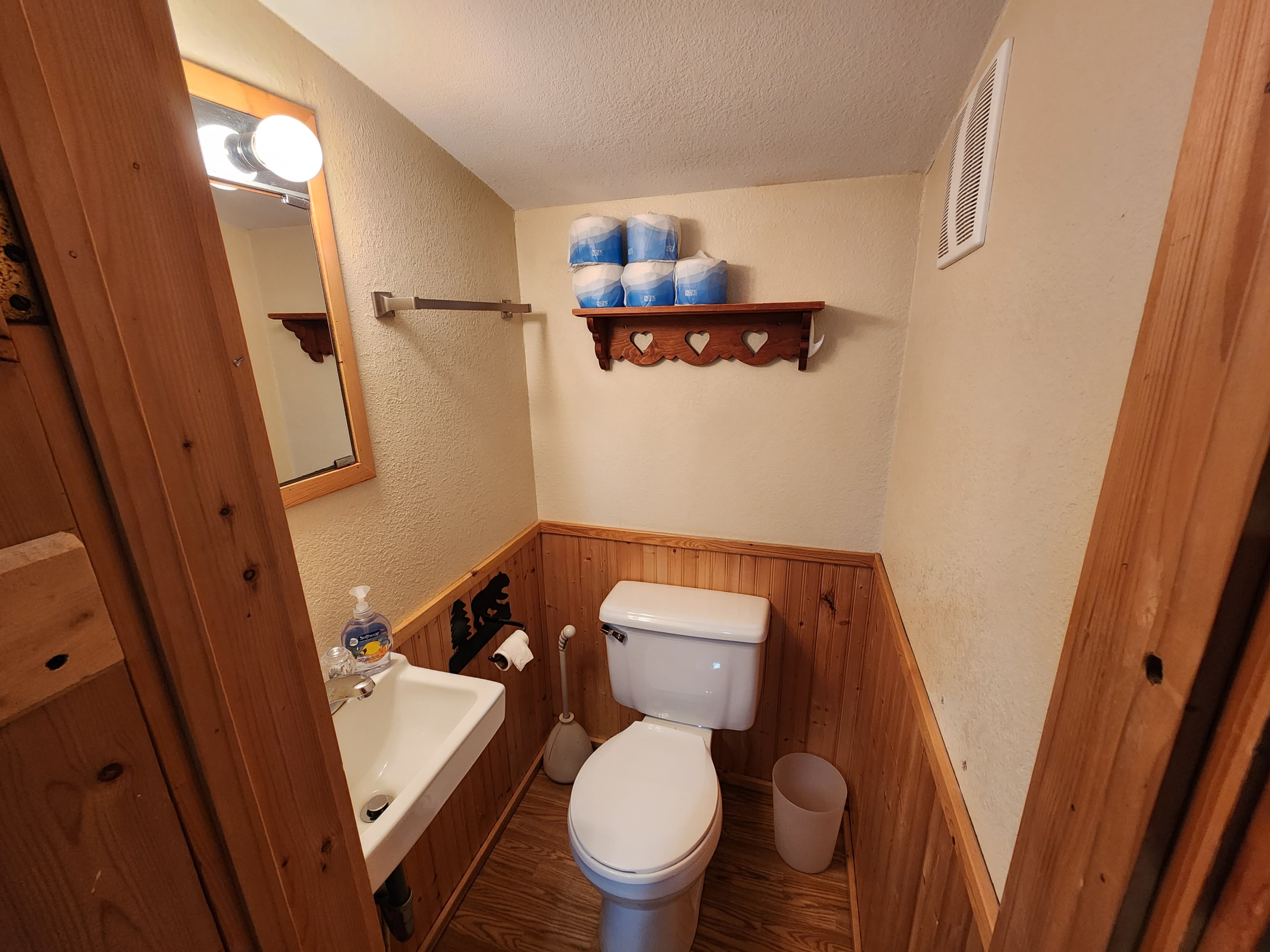 A small bathroom featuring a toilet, sink, mirror, and shelves with toilet paper.