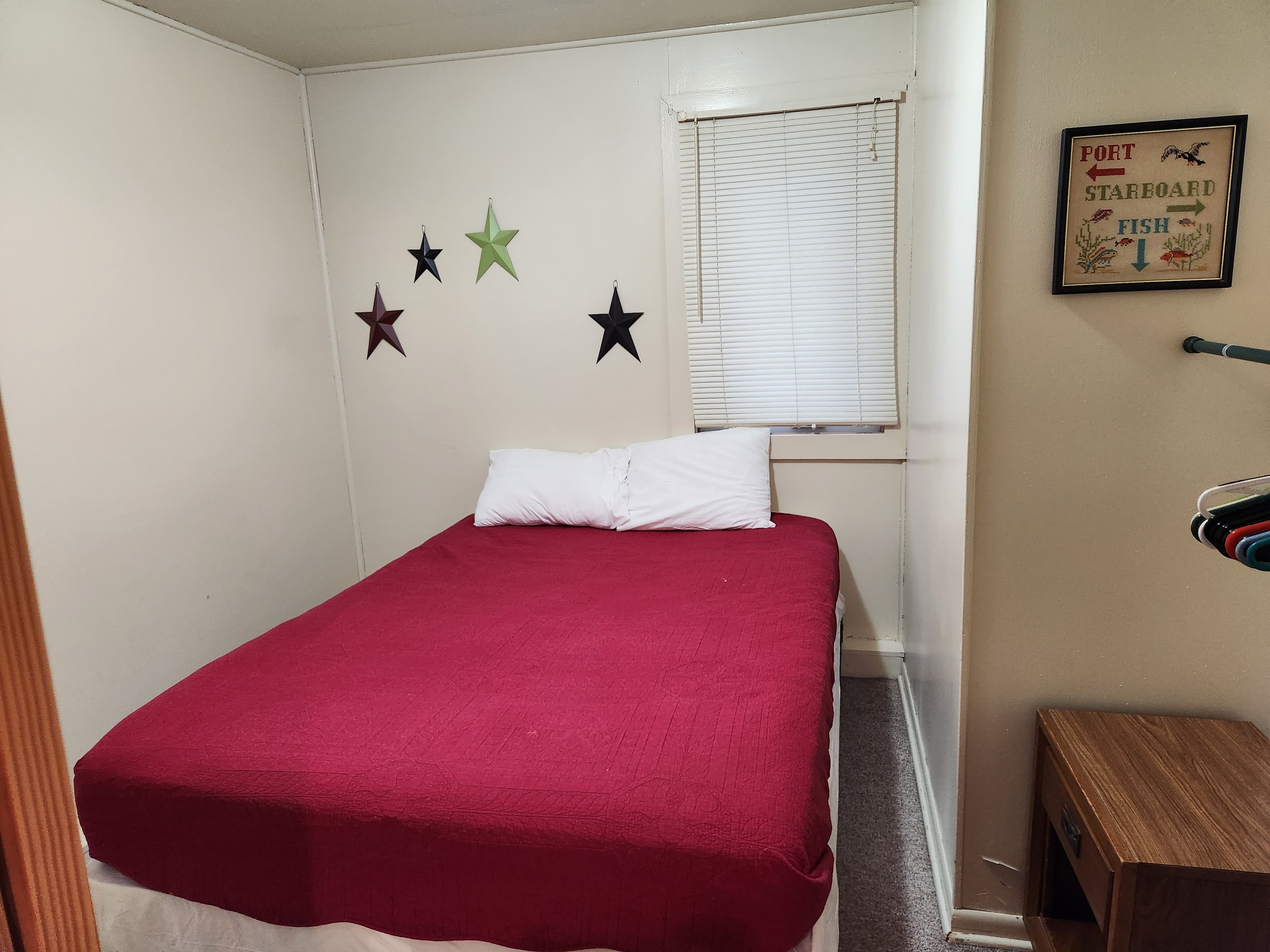 A small bedroom featuring a red bedspread, two pillows, decorative star wall art, and a window with blinds.