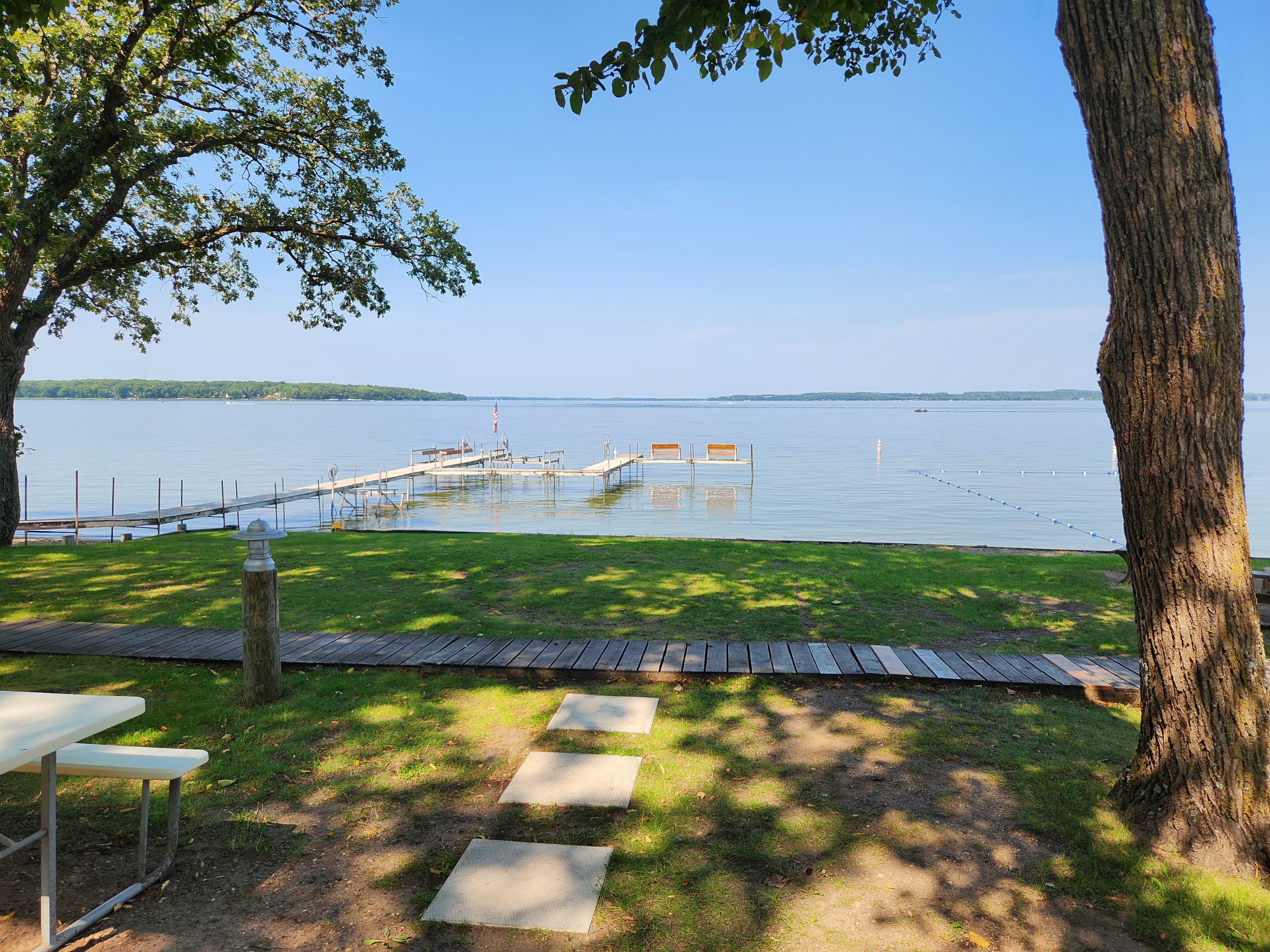 A serene lakeside view with a dock and wooden pathway framed by trees on a sunny day.