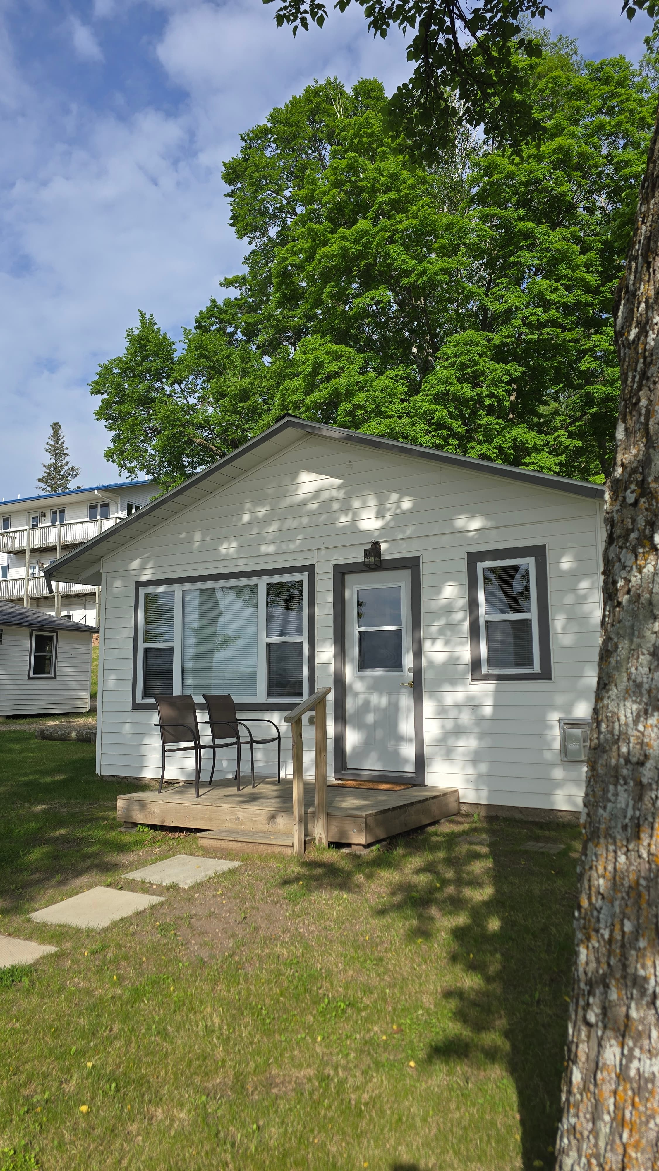 A small white cottage with a wooden deck and chairs, surrounded by green trees.