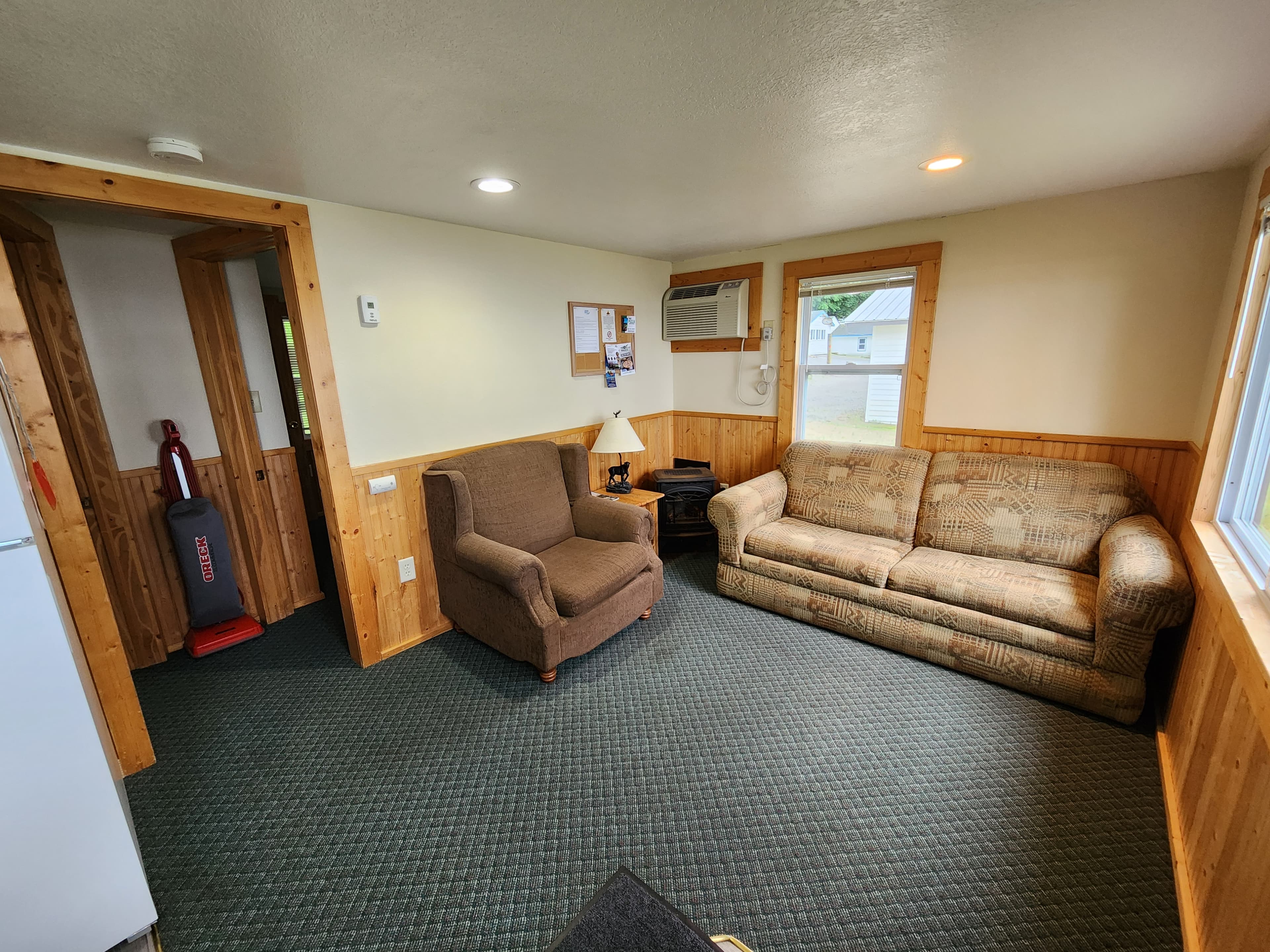A cozy living room featuring two chairs, a sofa, a lamp, and an air conditioner in a wood-paneled space.