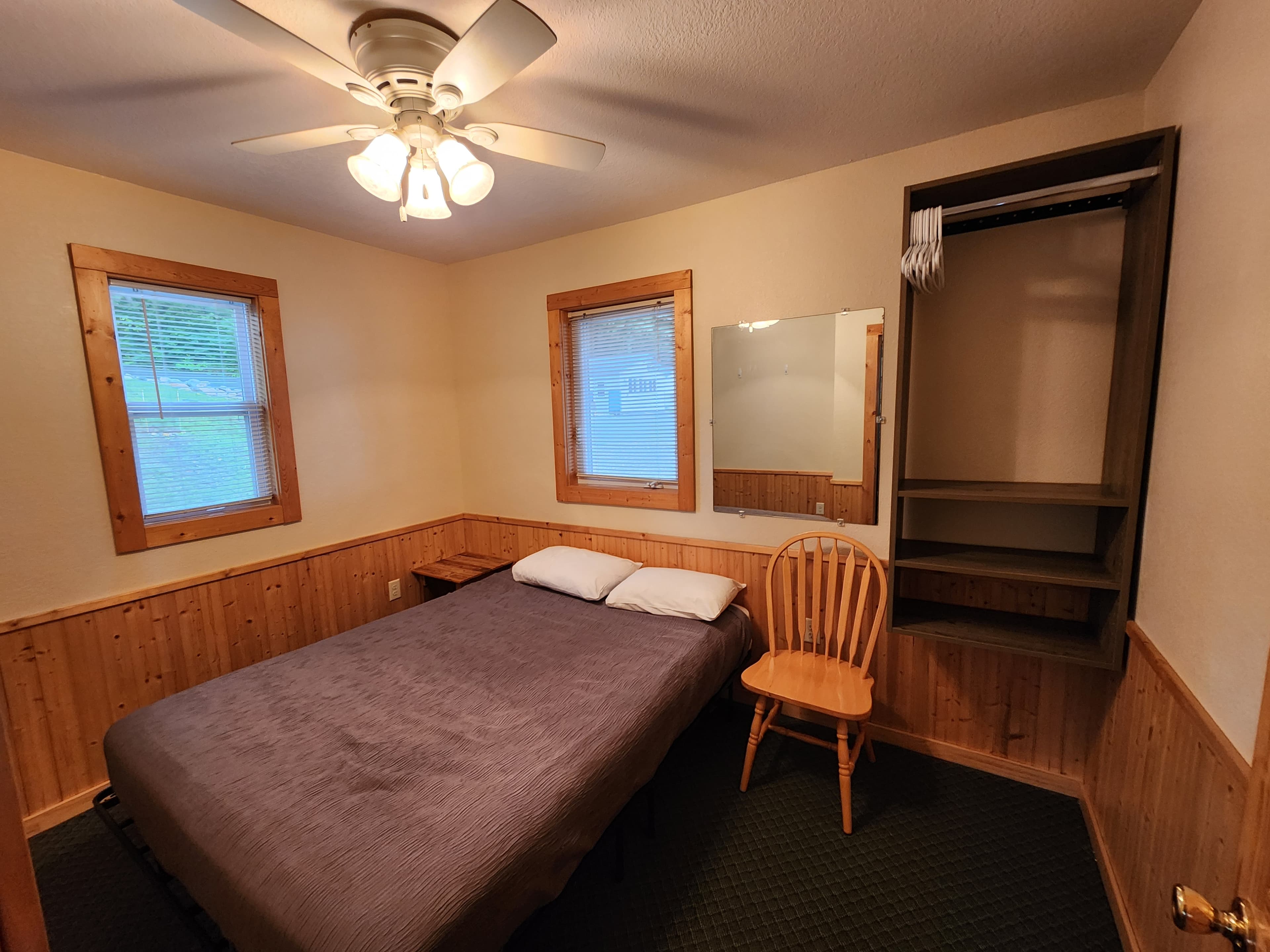 A simple bedroom featuring a bed, chair, mirror, and empty closet with wooden accents and natural light.