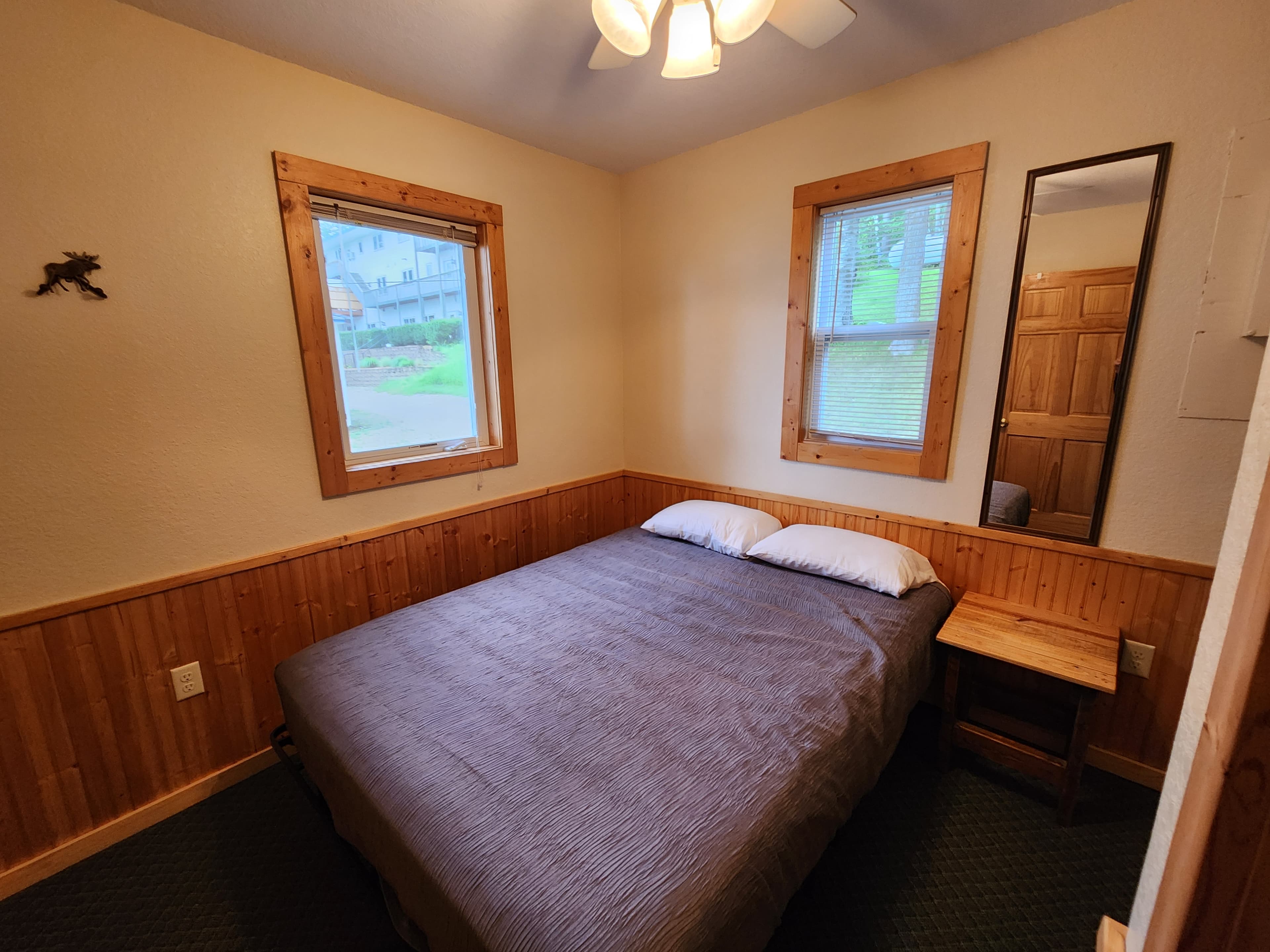 A cozy bedroom featuring a double bed, two windows, a small wooden table, and a mirror.