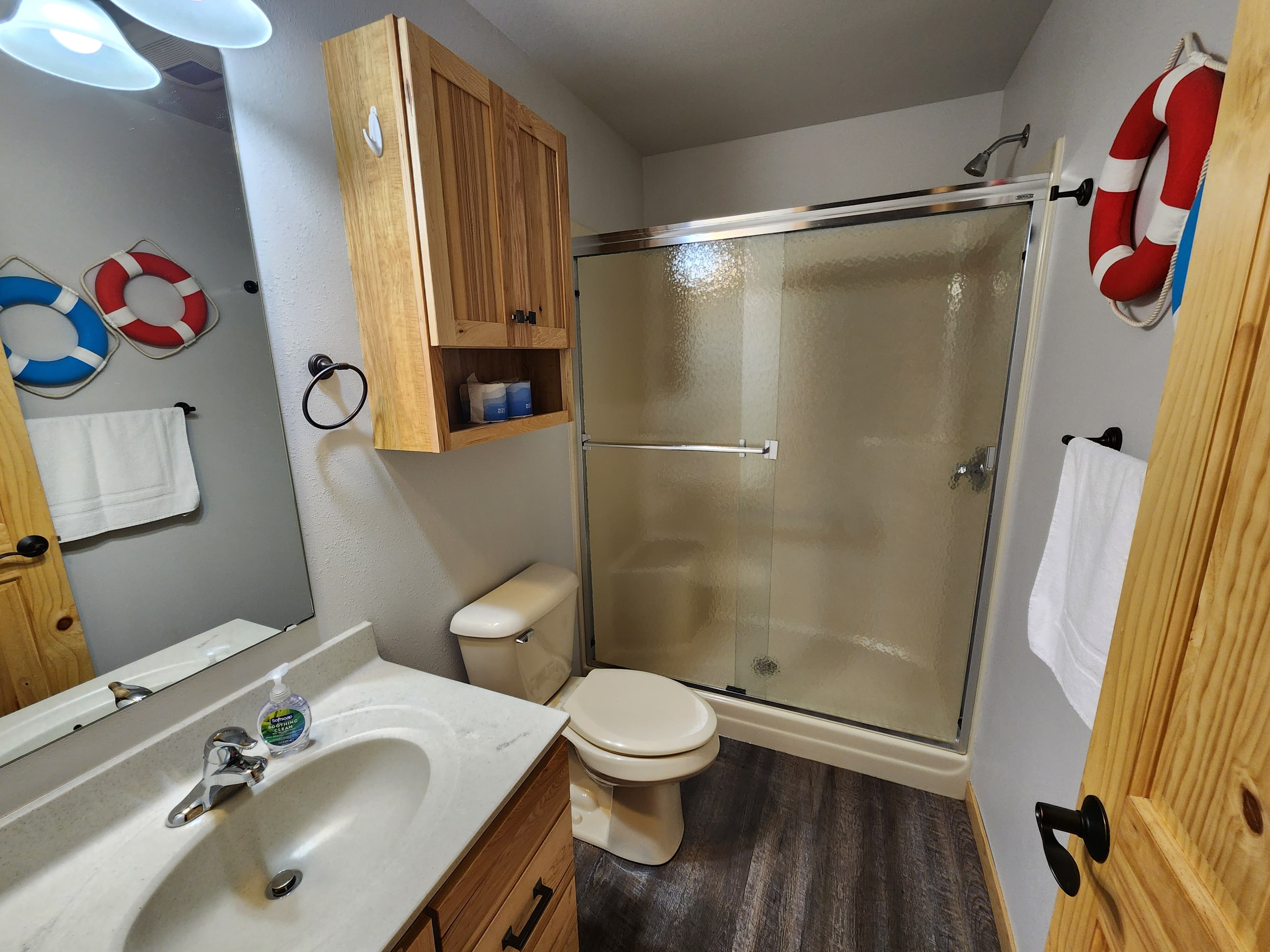 A light-colored bathroom featuring a shower, toilet, sink, and wooden cabinetry with life preservers on the walls.