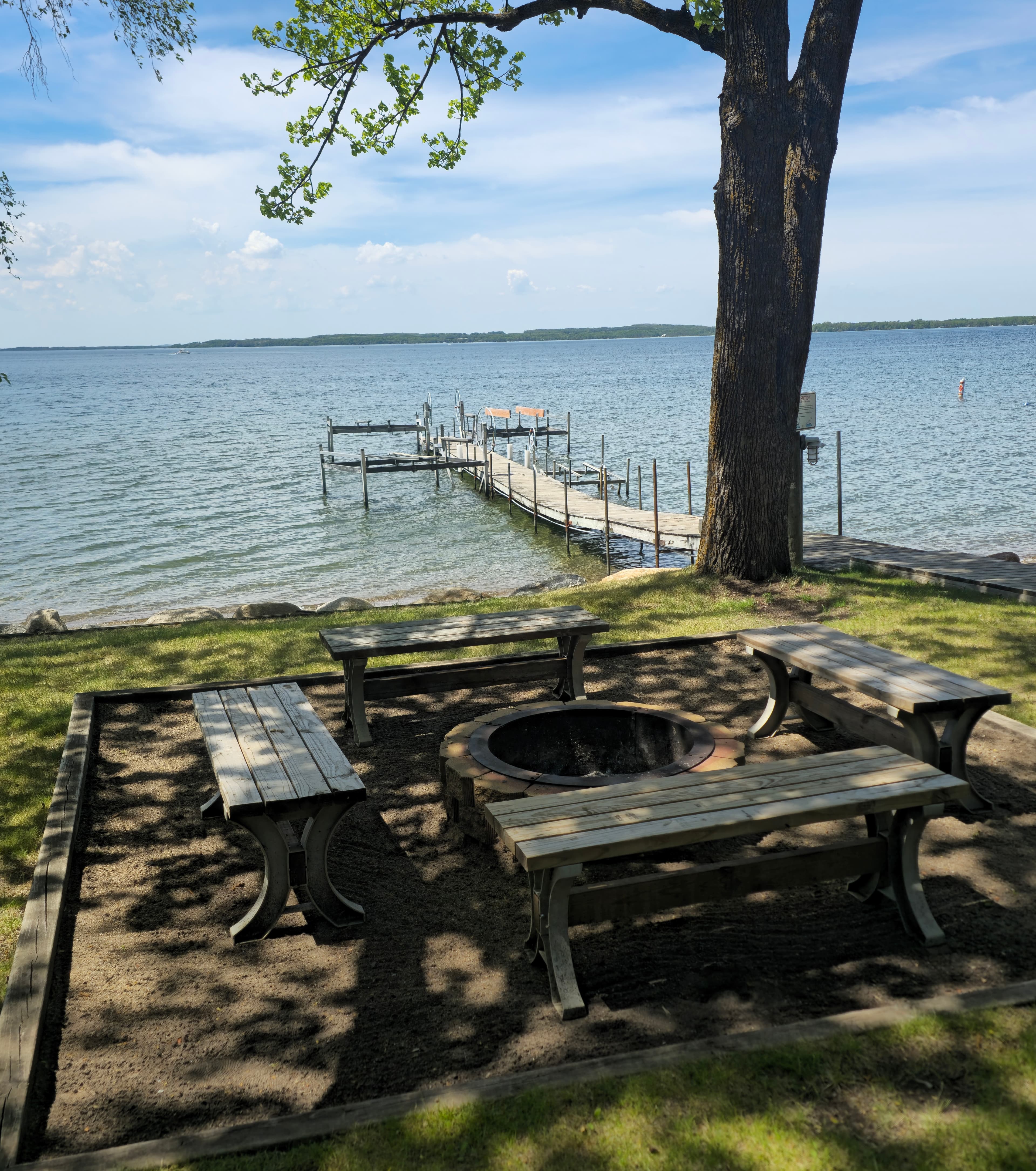 A lakeside scene featuring a wooden dock, a fire pit, and picnic tables under a tree.