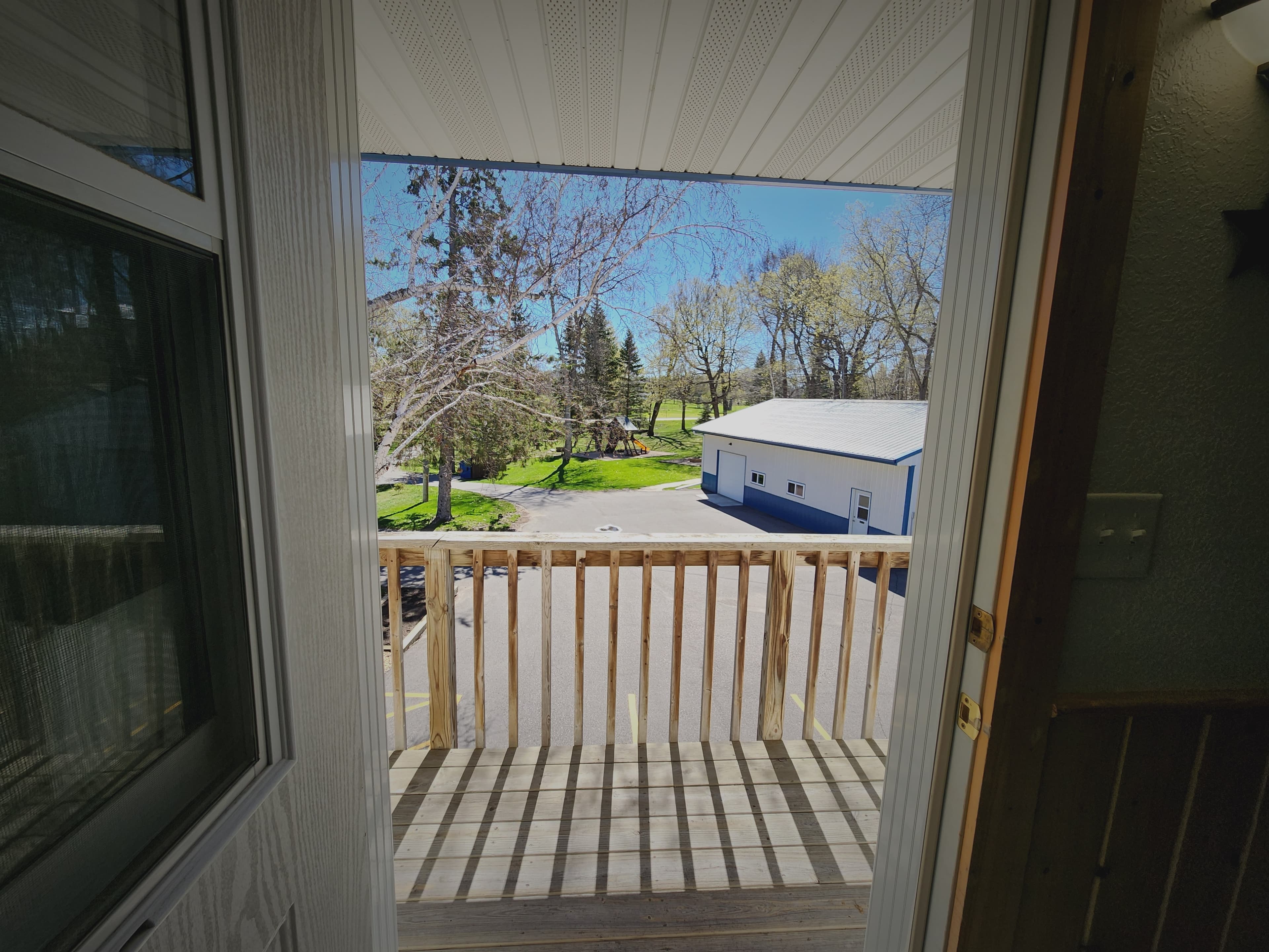 View from an open door onto a sunny balcony overlooking trees and a driveway.