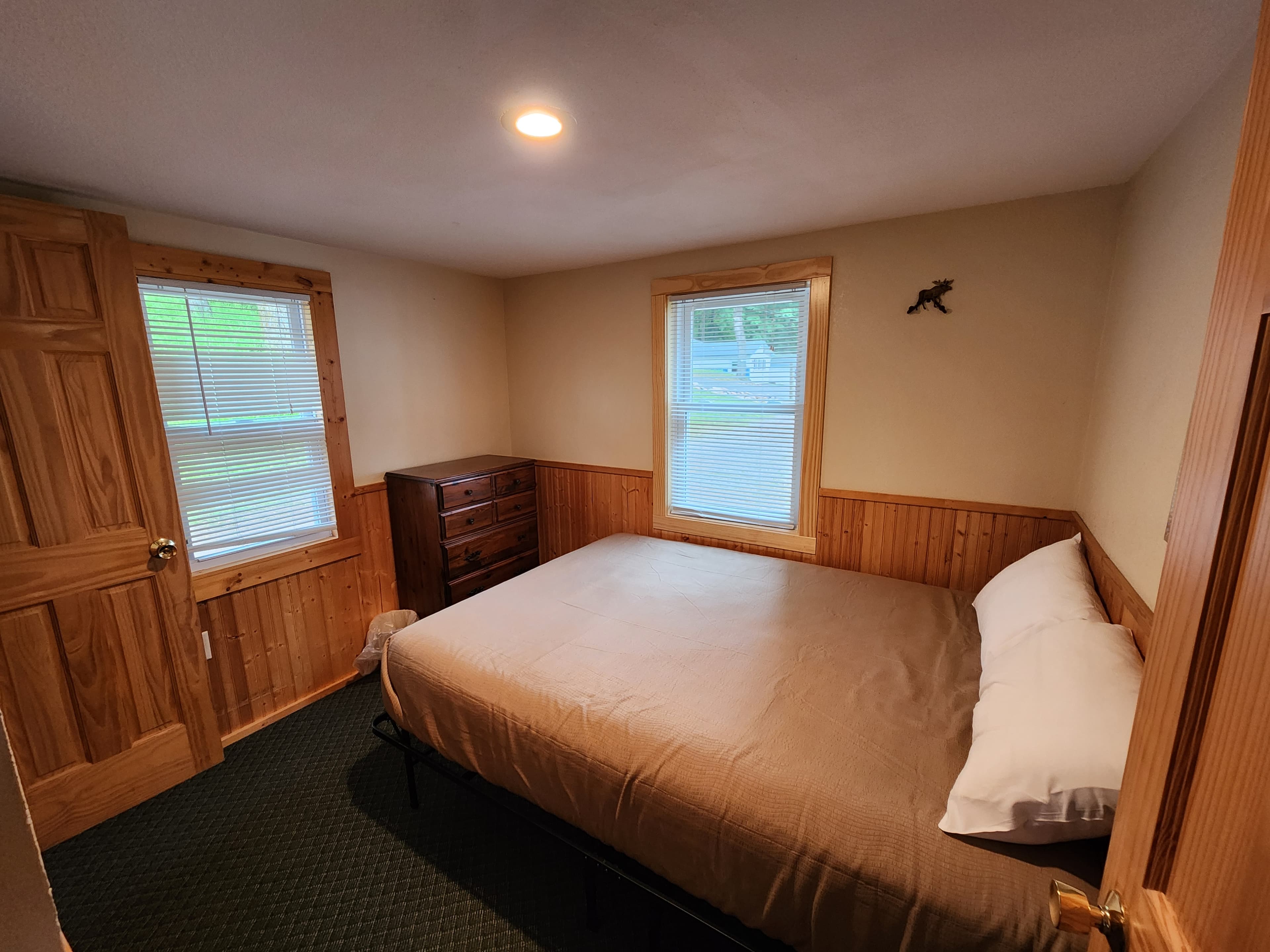 A cozy bedroom featuring a queen-sized bed, wooden furniture, and two windows with blinds.