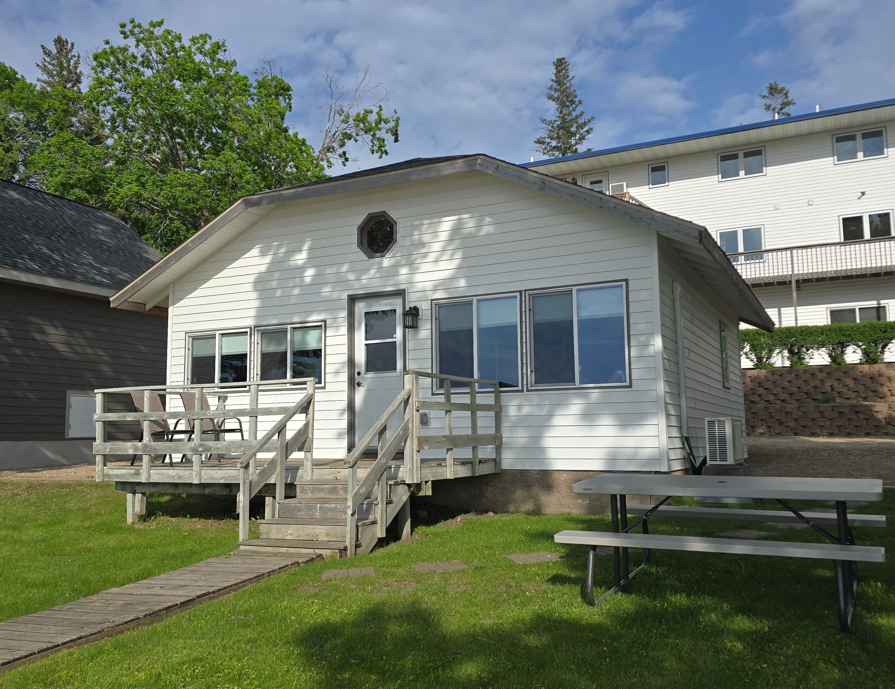 A white cottage with a wooden deck and picnic table on a grassy area.