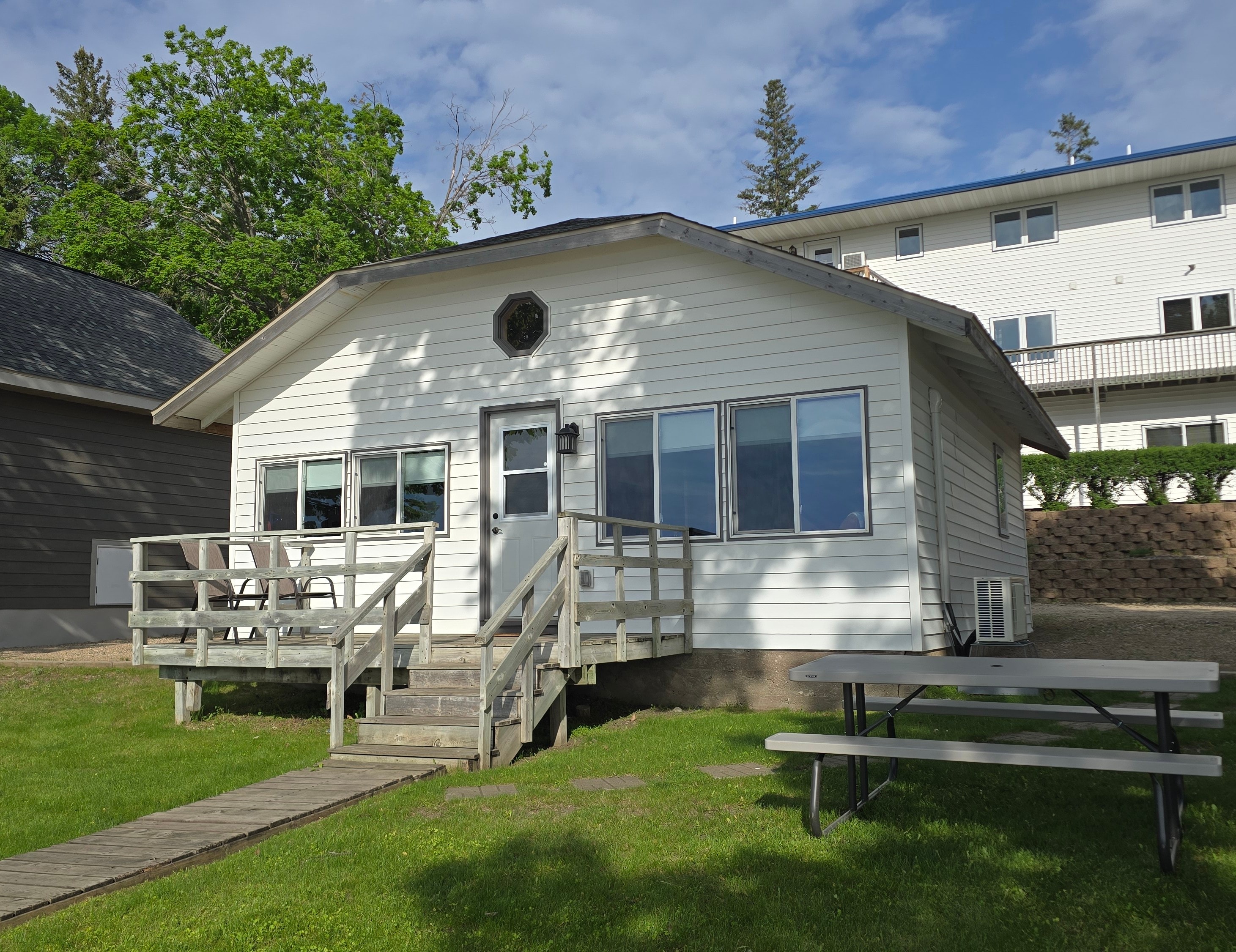 A white cottage with a wooden deck and picnic table on a grassy area.