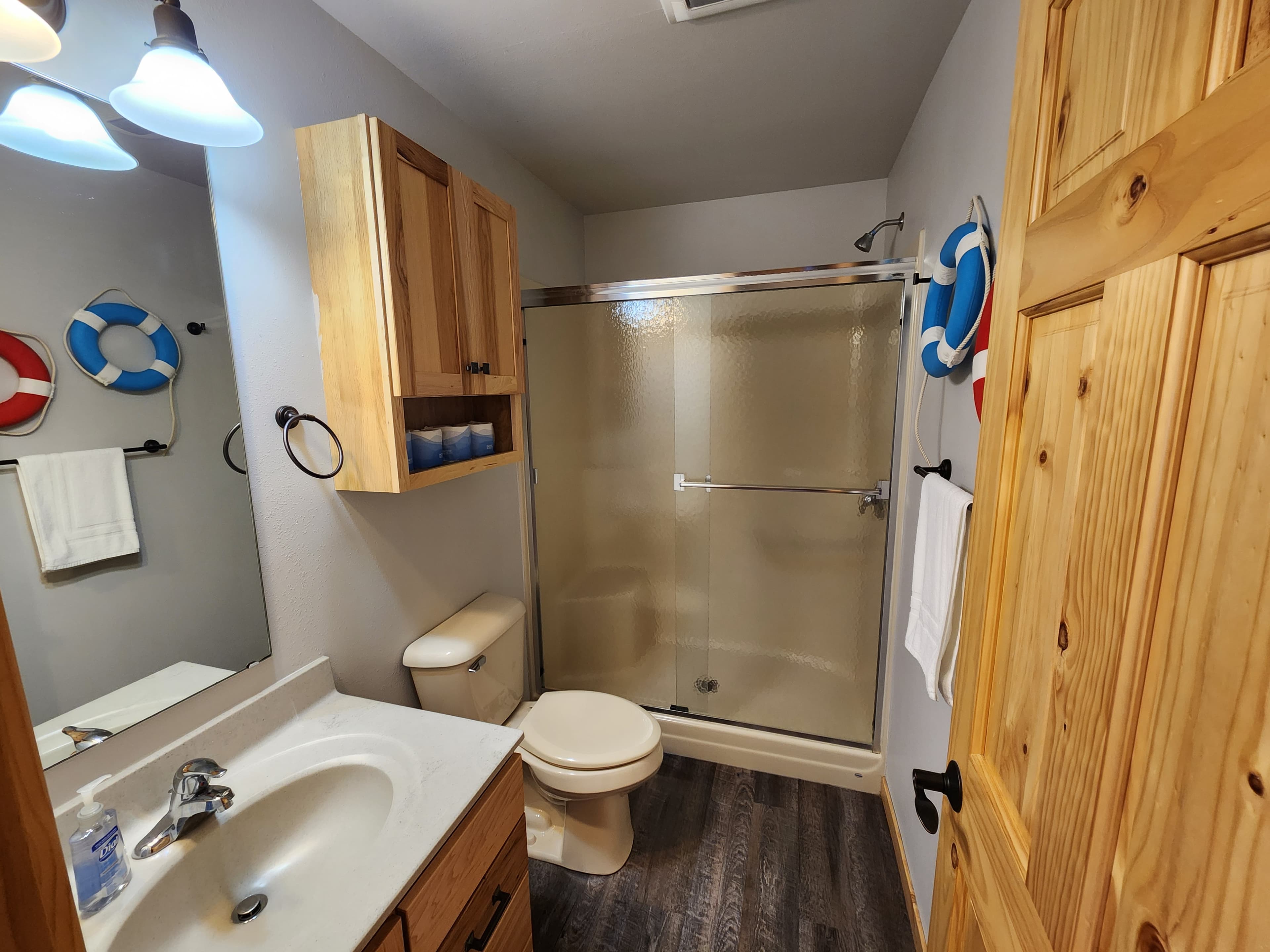 A bright bathroom featuring wooden cabinets, a shower, toilet, and nautical-themed decor.