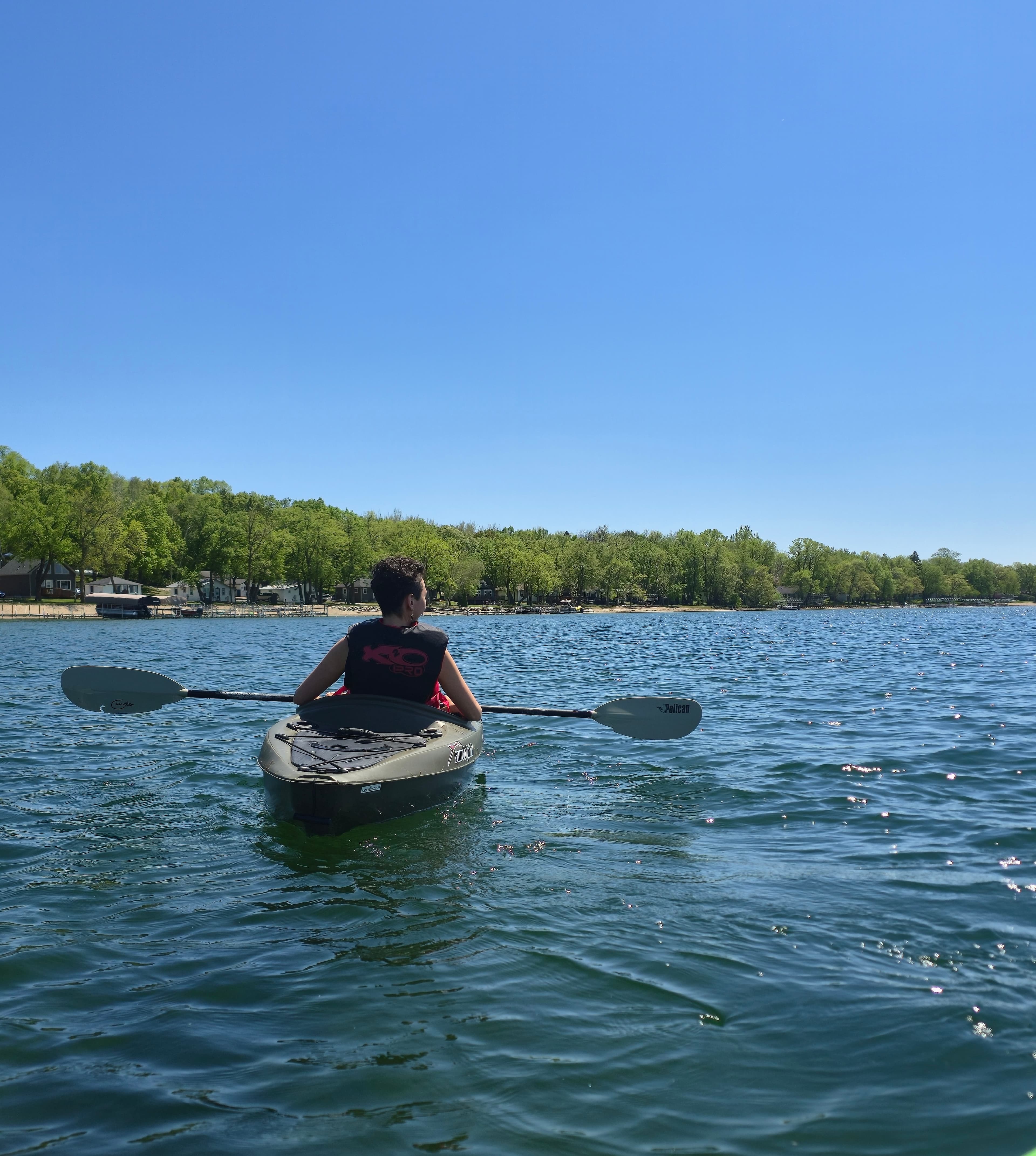 A person in a kayak paddles on a calm lake under a clear blue sky.