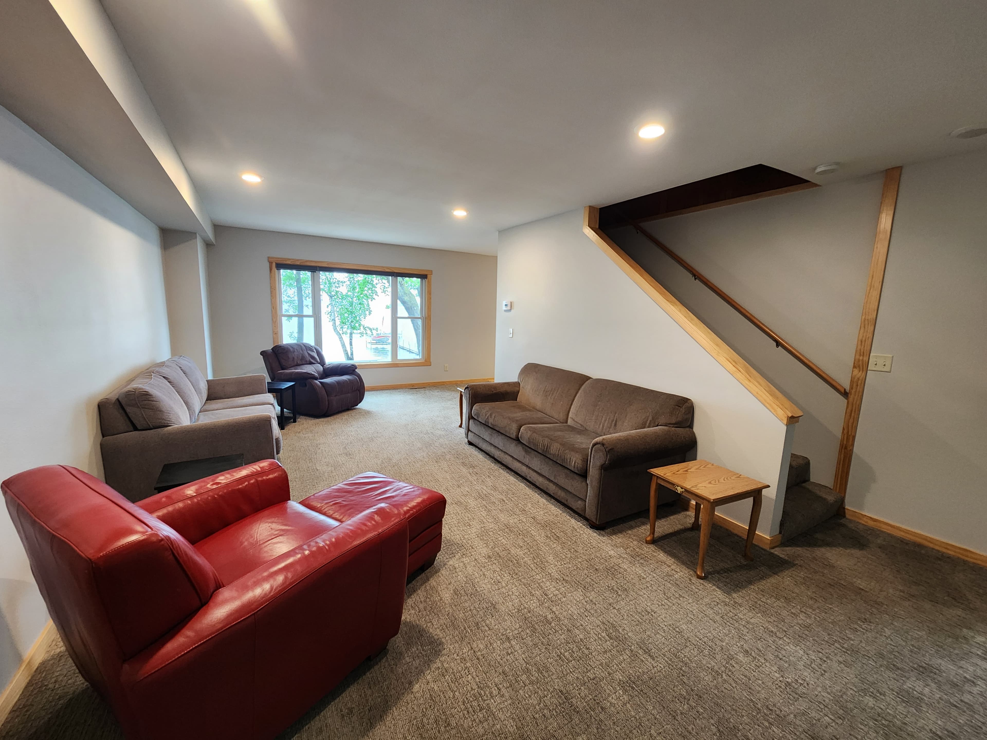 A spacious living room featuring a mix of seating options, including red and gray sofas, with a staircase in the corner and a large window letting in natural light.
