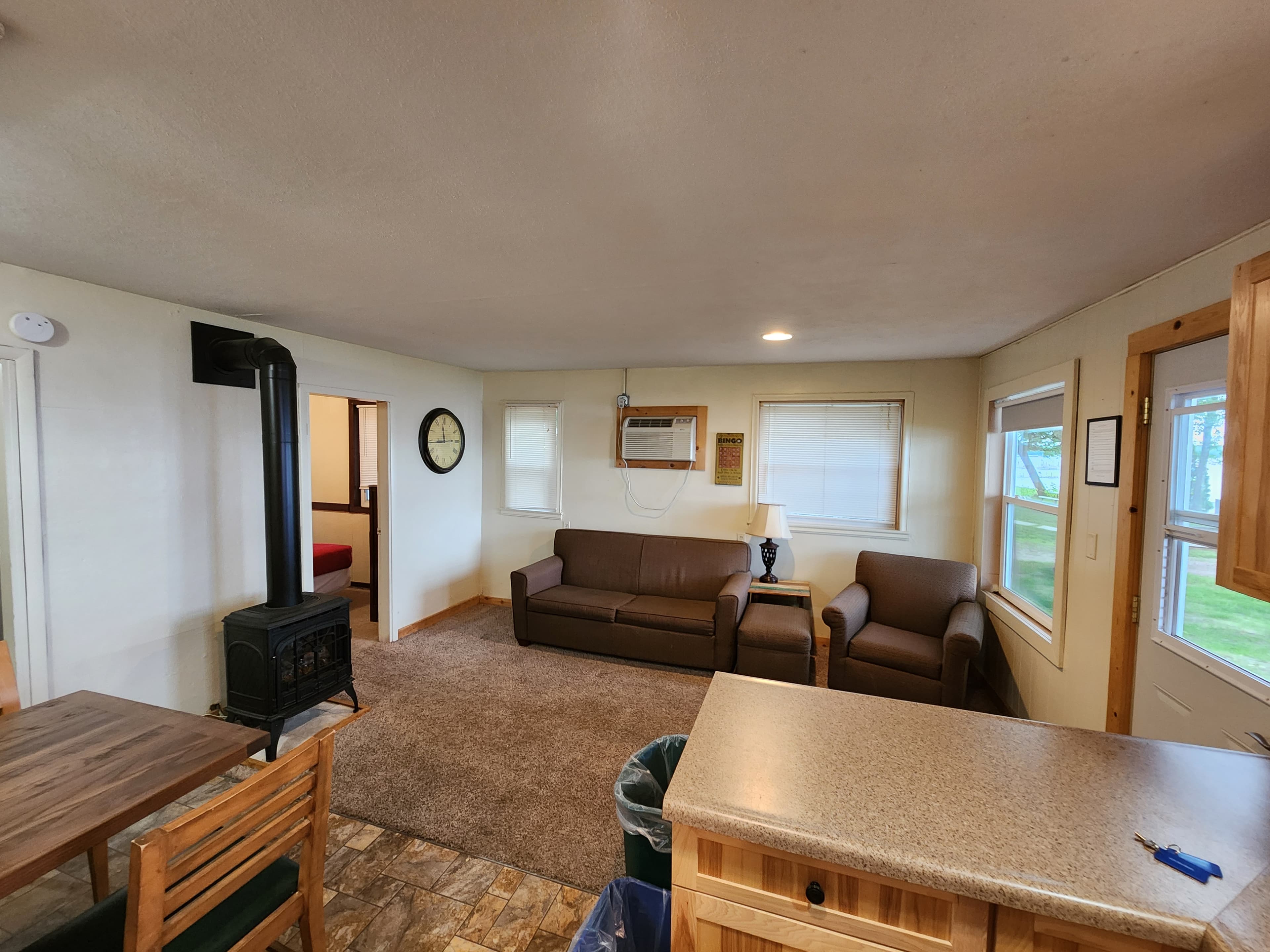 A cozy living area featuring a brown sofa, two chairs, a dining table, and a wood stove, with windows providing natural light.