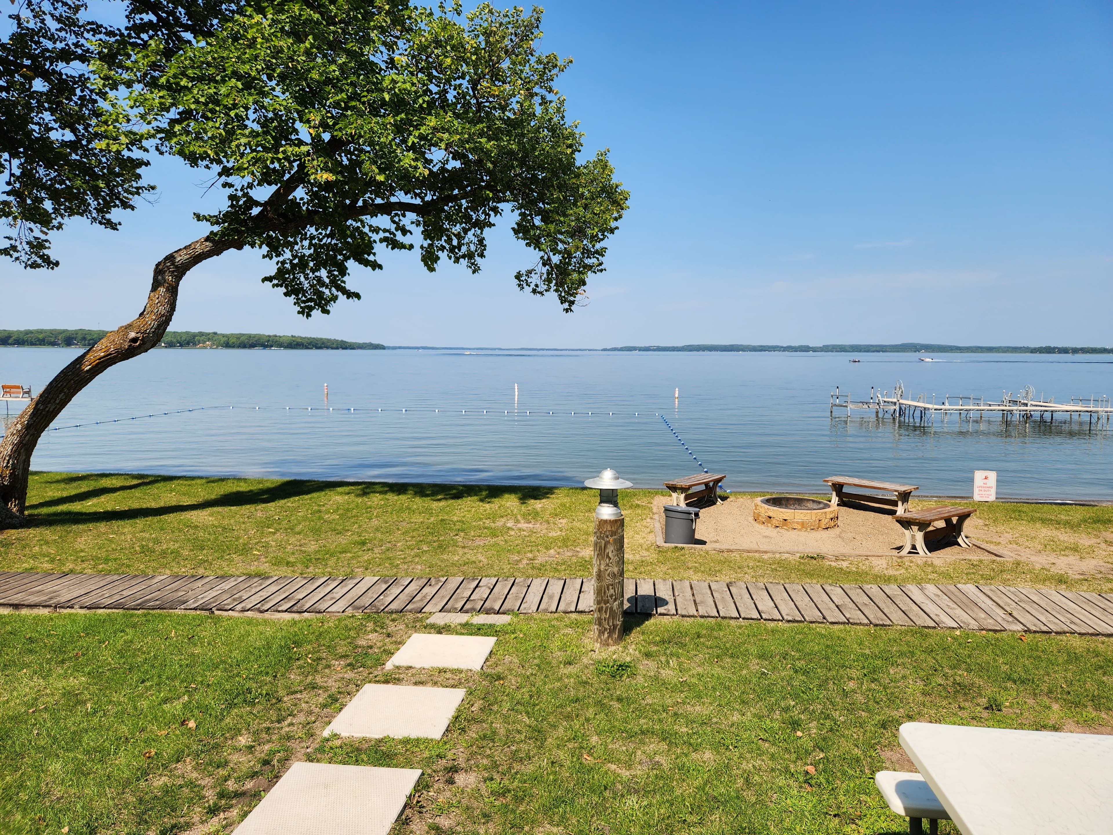 A serene lakeside view with a grassy shore, wooden seating, and a clear blue sky.
