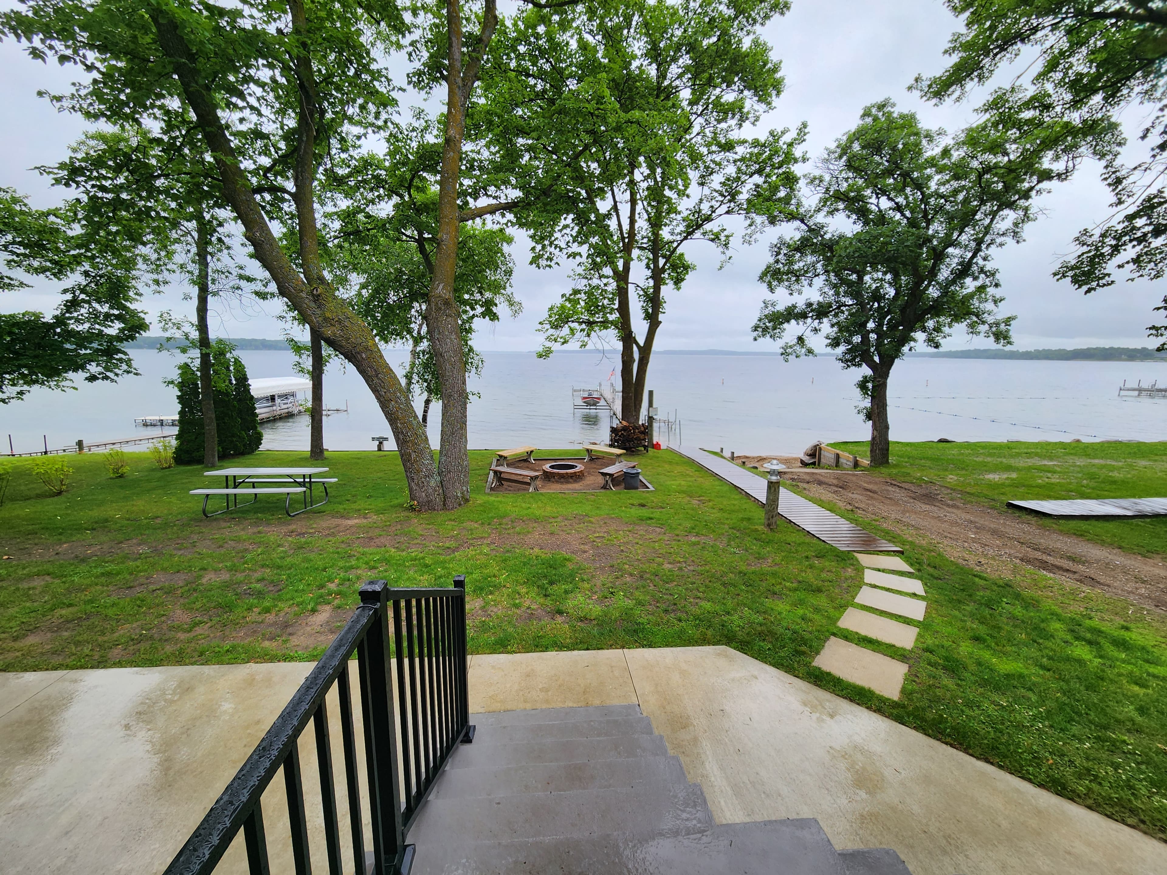 A grassy area with trees and a lake view, featuring a fire pit, picnic table, and steps leading down.